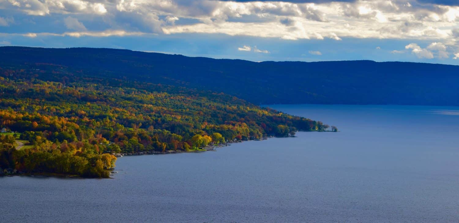 A scenic view of a lake bordered by colorful autumn foliage and rolling hills under a cloudy sky.